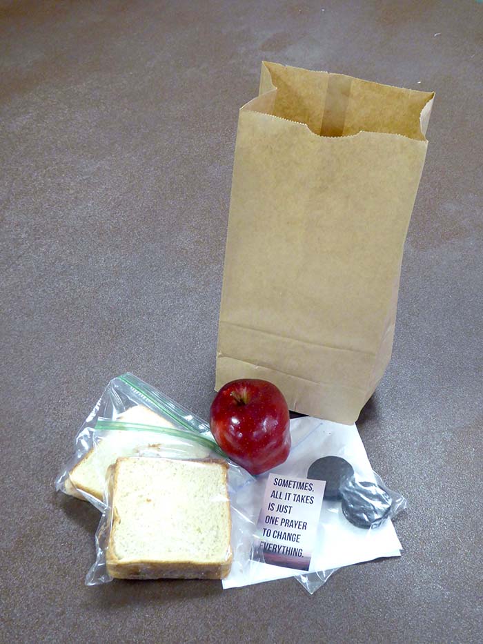 Each bag contains two peanut butter and jelly sandwiches, an apple, cookies, prayer card, bottle of water, and napkins.